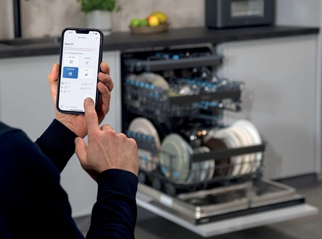 Vestavná myčka Haier Xynergy: Mimořádná úspora s tepelným čerpadlem ruce držící mobil a aplikací hon, v pozadí je vestavná myčka Haier Xynergy